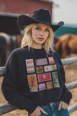 Woman wearing a black cowboy hat and a black sweatshirt with colorful graphics, standing in front of a wooden fence with horses in the background.