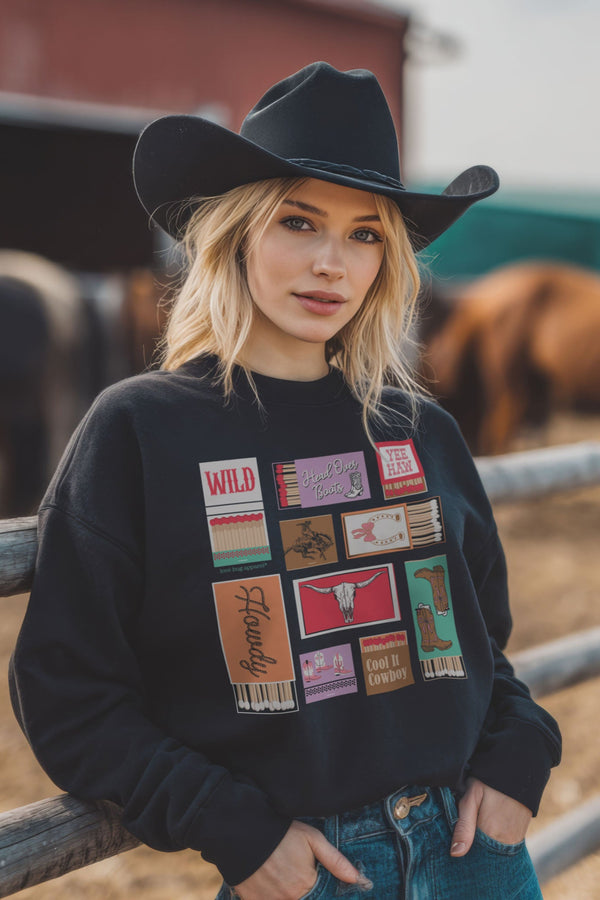 Woman wearing a black cowboy hat and a black sweatshirt with colorful graphics, standing in front of a wooden fence with horses in the background.