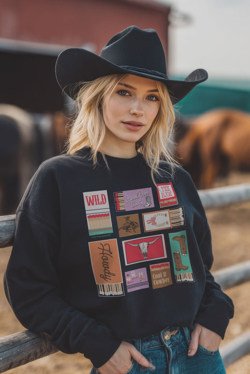 Woman wearing a black cowboy hat and a black sweatshirt with colorful graphics, standing in front of a wooden fence with horses in the background.