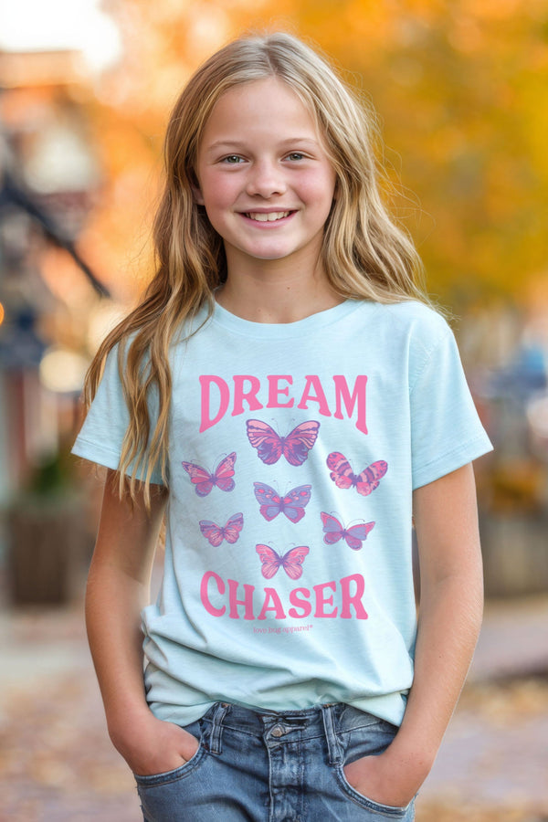 Young girl wearing a light blue t-shirt with pink butterfly design and text, standing outdoors with blurred background