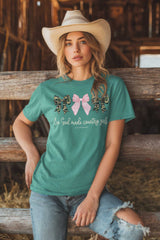 Woman wearing a teal t-shirt with a graphic design, sitting on a wooden fence in a barn.