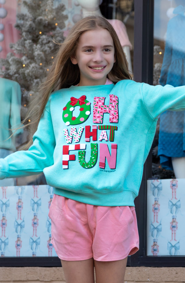 Young girl wearing a light blue sweatshirt with colorful text and pink shorts, standing in front of a store window display.