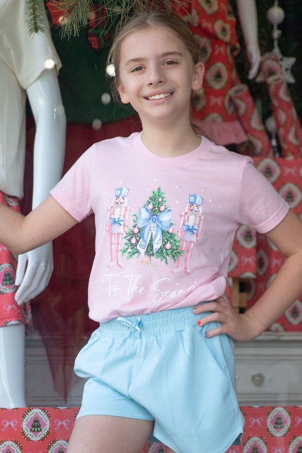 Young girl wearing a pink t-shirt with a Christmas Nutcrackers design and light blue shorts, standing in front of a decorated Christmas tree.