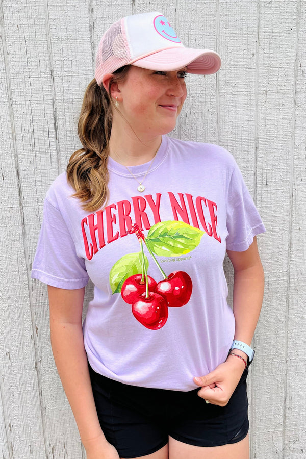 Person wearing a 'Cherry Nice' t-shirt and pink cap against a wooden background