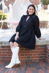 Woman in a black dress and white boots sitting on a brick ledge with a fountain in the background.