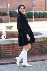 Woman in a black dress and white boots standing in front of a brick wall with greenery.
