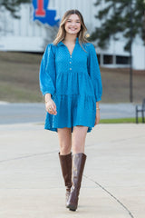 Woman in a blue dress and brown boots standing on a campus with a building and 'TL' logo in the background.