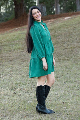 Woman in a green dress and black boots standing on a grassy hill with trees in the background