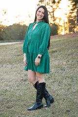 Woman in a green dress and black boots standing in a grassy field with trees in the background.
