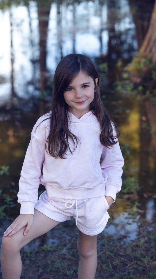 Young girl in a pink hoodie and shorts sitting by a forest stream.