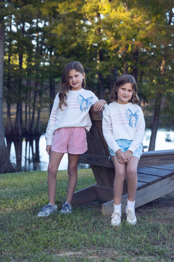 Two young girls sitting on a wooden bench by a lake with trees in the background.