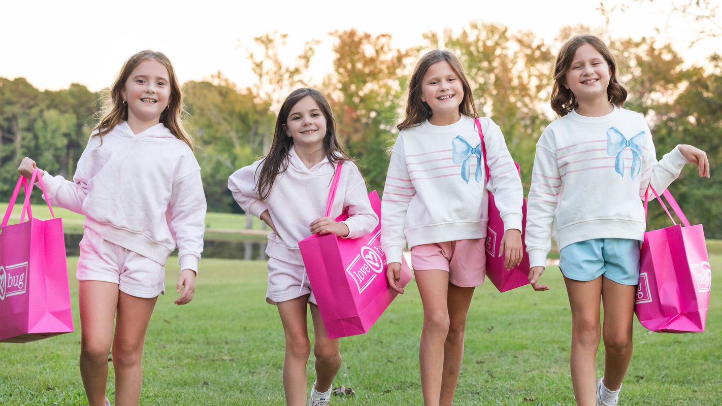 Four young girls in matching outfits with pink bags standing in a park.