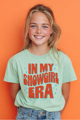 Young girl wearing a t-shirt with 'IN MY SHOWGIRL ERA' text against an orange background
