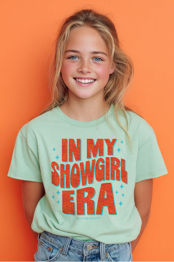 Young girl wearing a t-shirt with 'IN MY SHOWGIRL ERA' text against an orange background